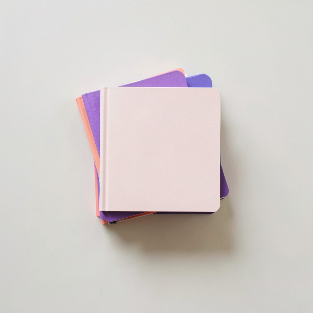 Overhead flat-lay photograph of a stack of four small notebooks with colored spines arranged on a pale surface, soft daylight, violet and coral accent colors, organized and nostalgic mood.