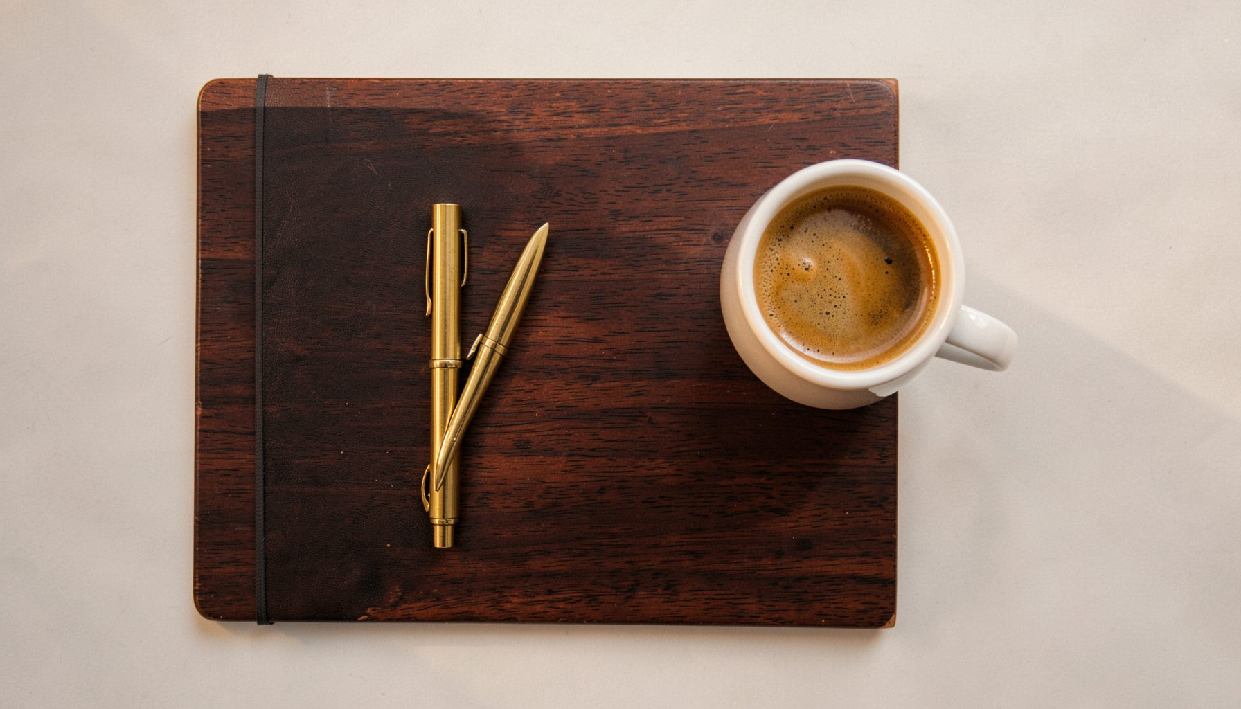 Overhead flat-lay photograph of a dark walnut desk with a brass pen, a small notebook, and a steaming espresso in a white ceramic cup, lit by warm morning light with soft shadows.