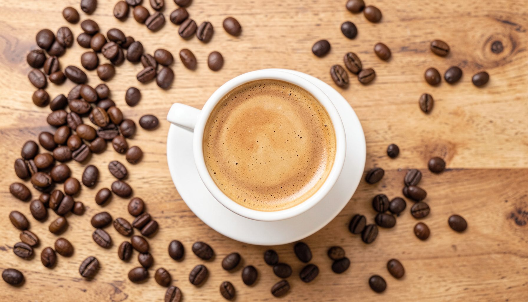 Overhead flat lay of a flat white in a white cup on a wooden table, surrounded by coffee beans, warm brown and cream tones, cozy cafe mood